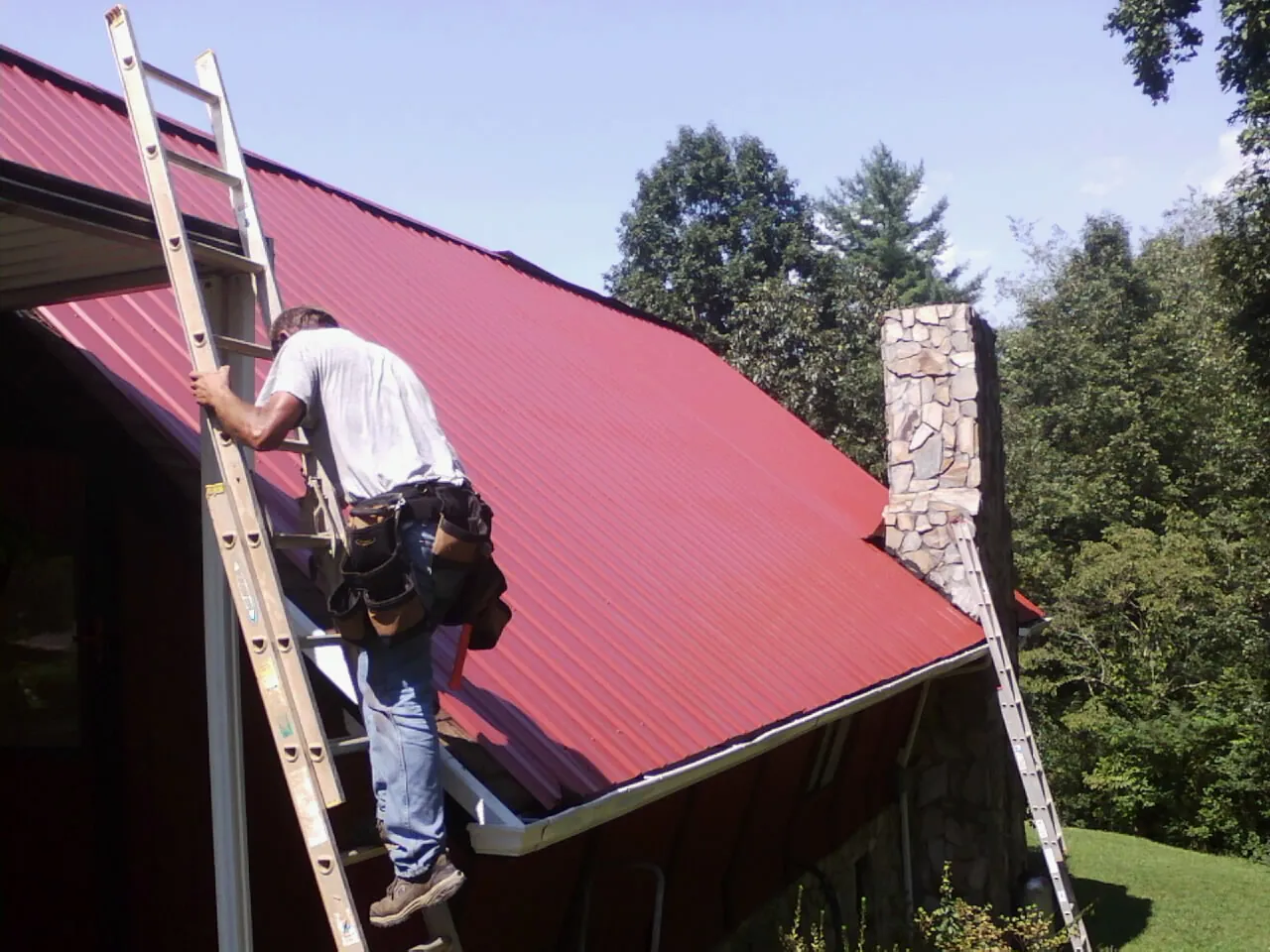 Roofer on a ladder beside a red metal roof and stone chimney — Soffit & Fascia Repair by Roofing Brothers in Paullina